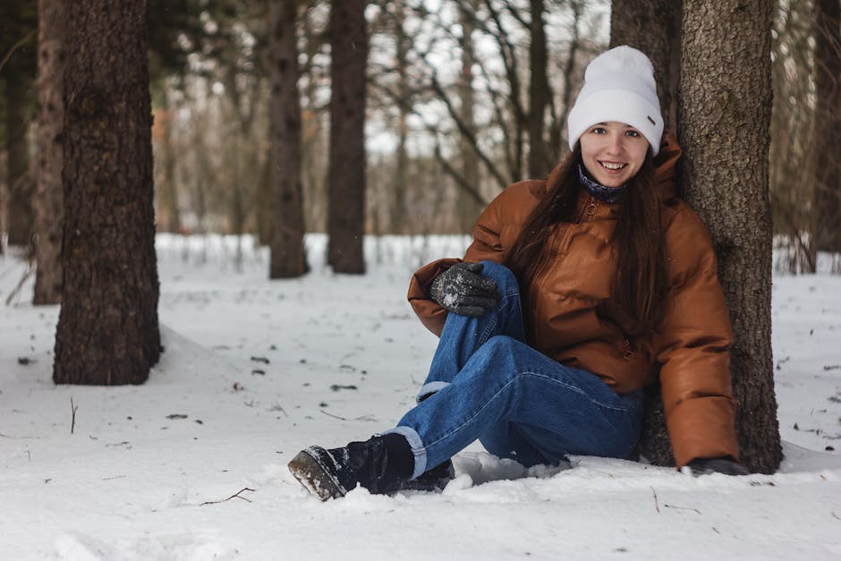 Battery Heated Clothing for Outdoor Activities - A woman wearing a beanie and winter clothes sits against a tree in a snow-covered forest, smiling warmly.