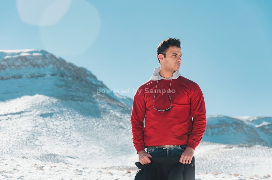 battery heated shirt outdoor activities - Man in red jacket standing in snowy landscape of Fordo, Iran, with mountainous backdrop.