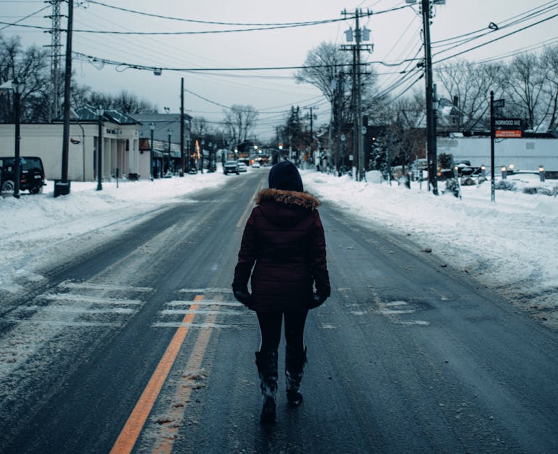 electric heated garments outdoor activities - A person in winter clothing walks down a snow-covered urban street during daylight.