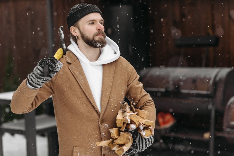heated wear outdoor activities - A bearded man wearing a beanie and brown coat chops wood outdoors during a snowfall.